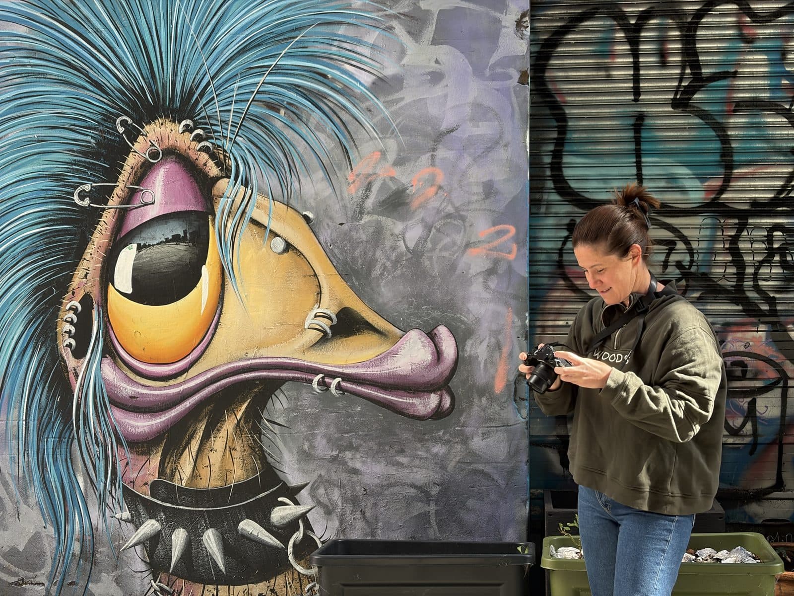 Eliza checking the back of her Fujifilm X-S20 beside a large blue-crested bird mural in a Melbourne laneway during a beginner photography course