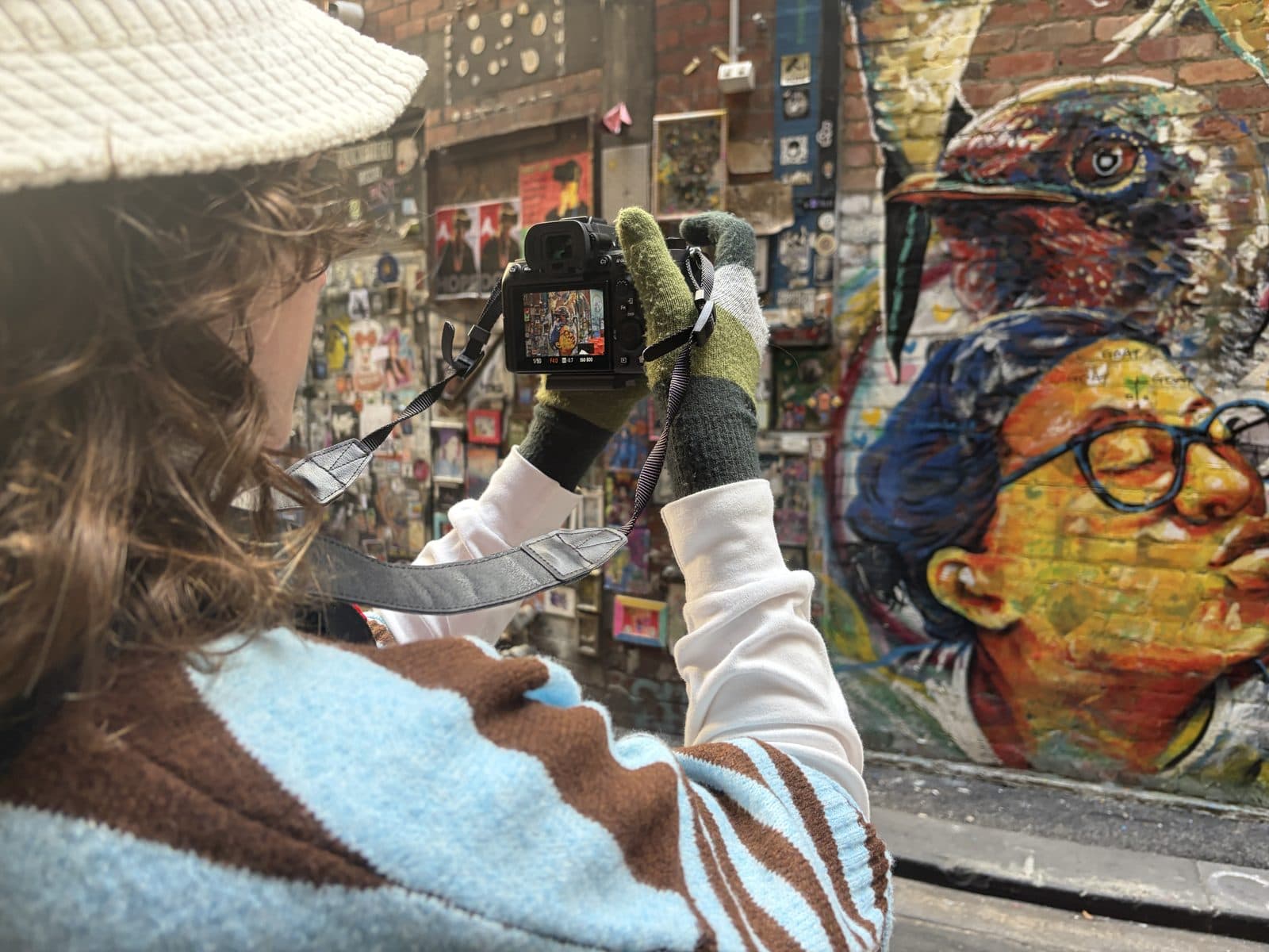 Julia's Sony A7 III rear screen showing manual mode settings 1/50 f4 ISO 800 while she photographs a street-art wall in a Melbourne laneway