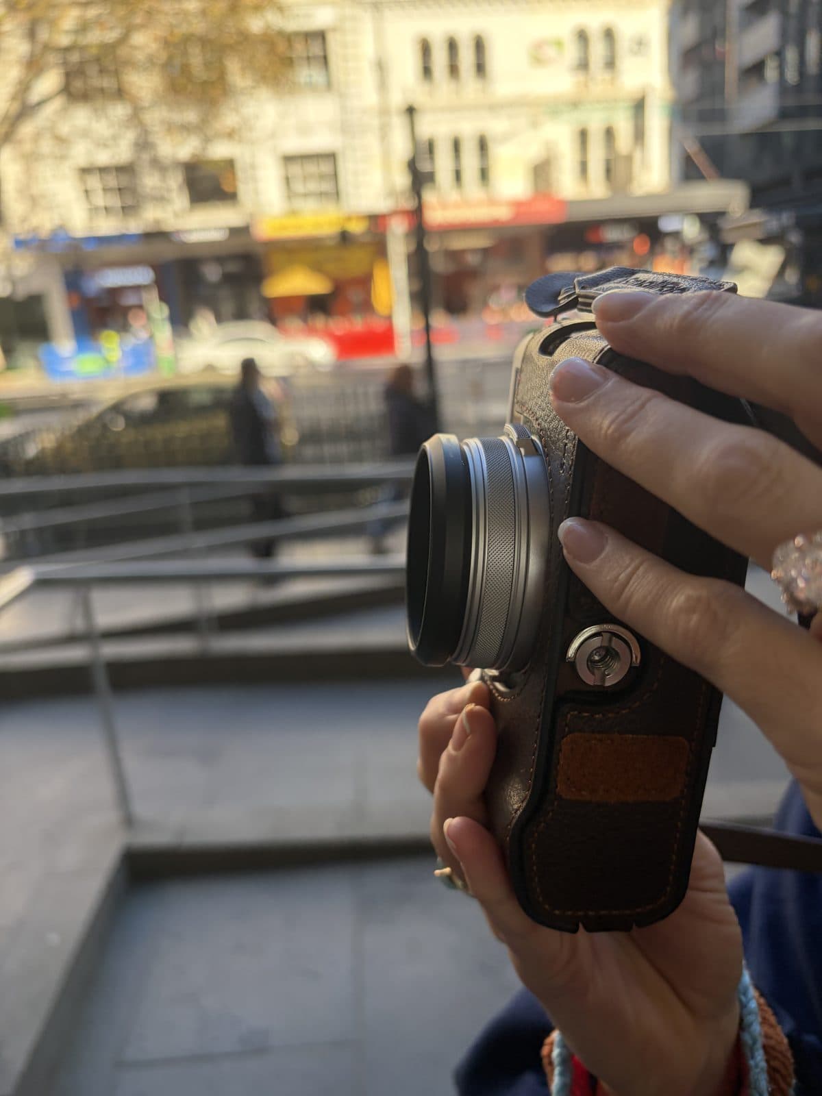 Close-up of a Fujifilm X100VI with a lens hood, brown leather half-case and SmallRig grip, city laneway behind